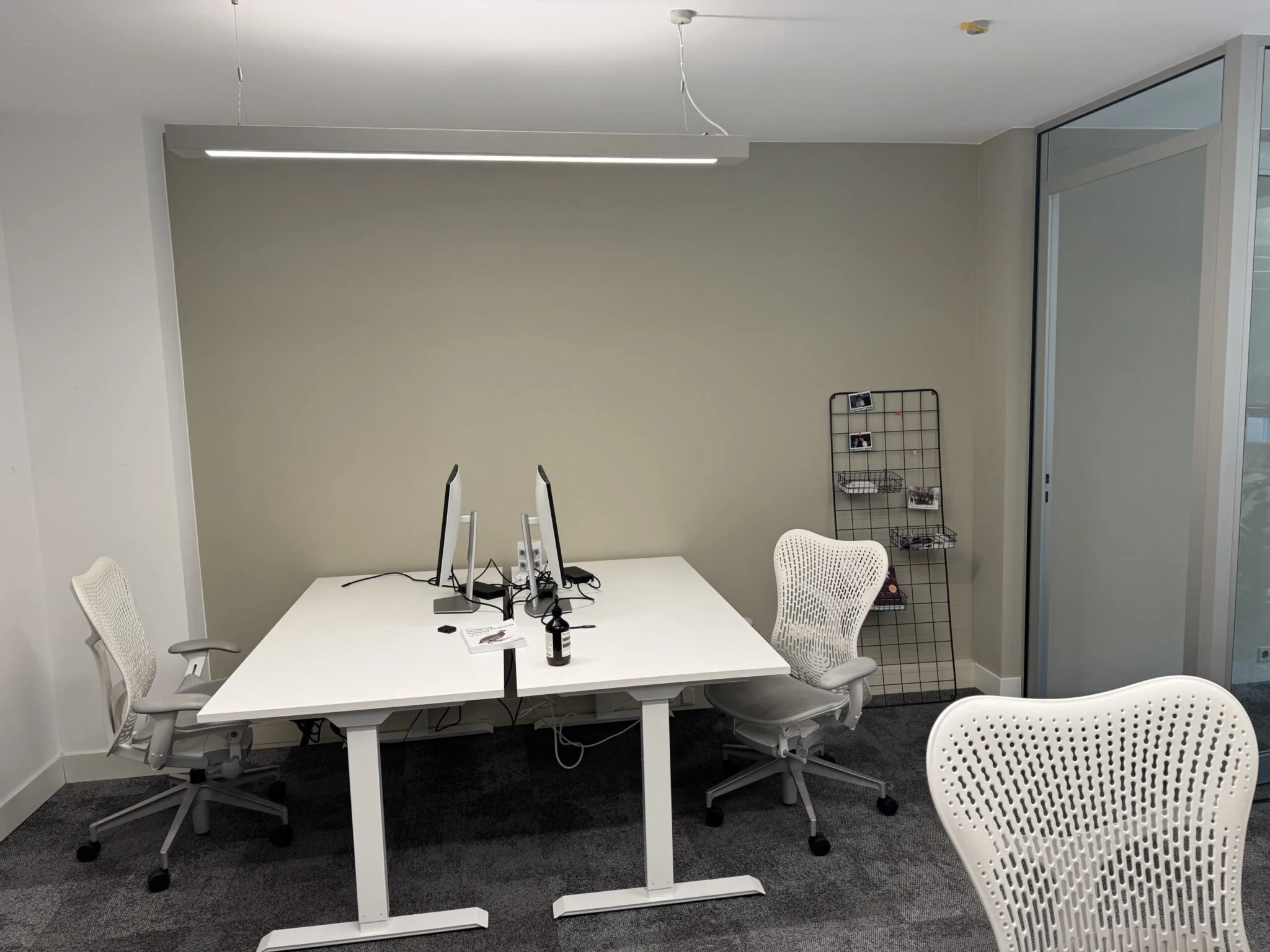 Moderne kantoorruimte met twee bureaus, ergonomische stoelen, computerschermen en een rek tegen de muur.