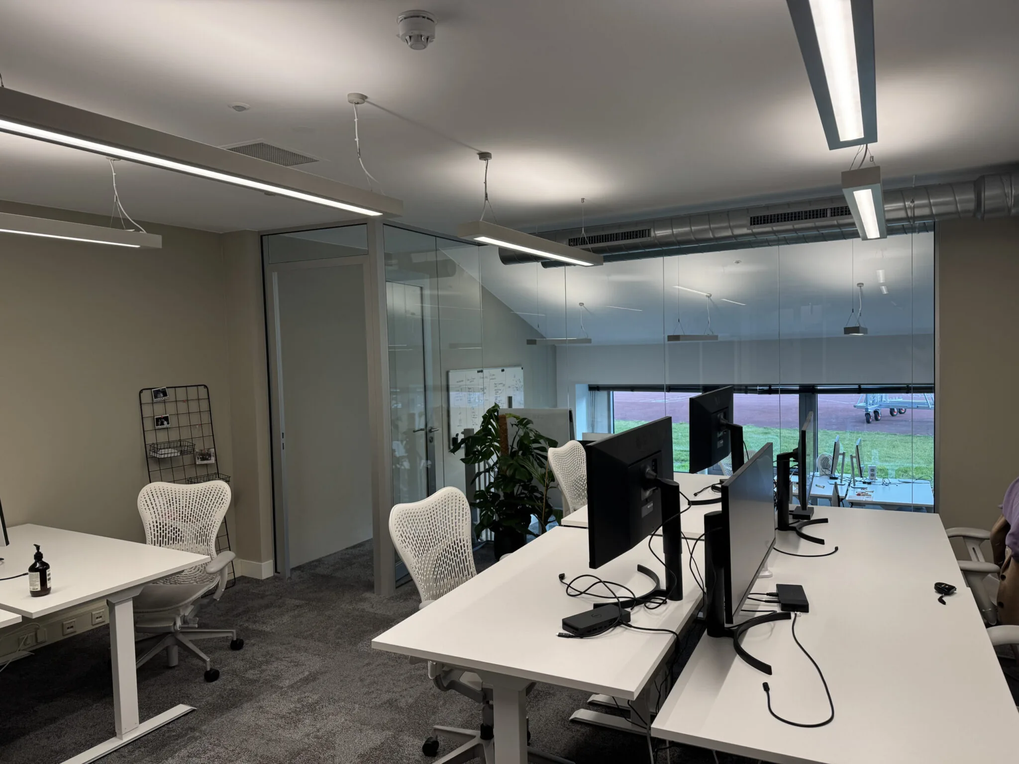 Moderne kantooruimte met witte bureaus, ergonomische stoelen en meerdere computerschermen, grenzend aan een glazen vergaderruimte.