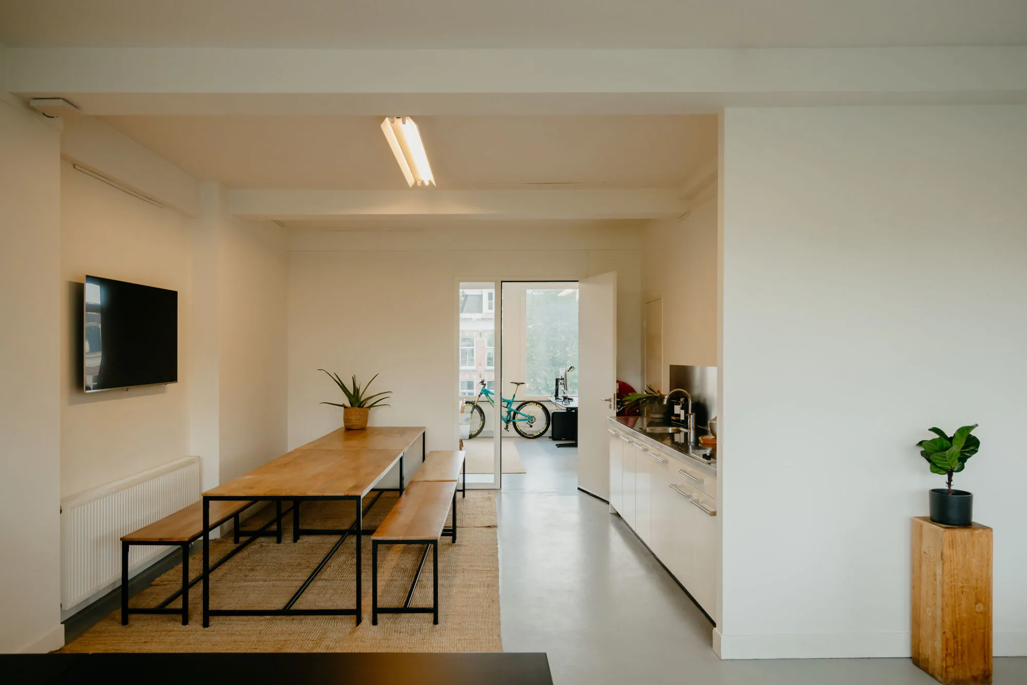 Een moderne eetkamer en keuken met een houten tafel en banken, een tv aan de muur, en een deuropening naar een kamer met een fiets.