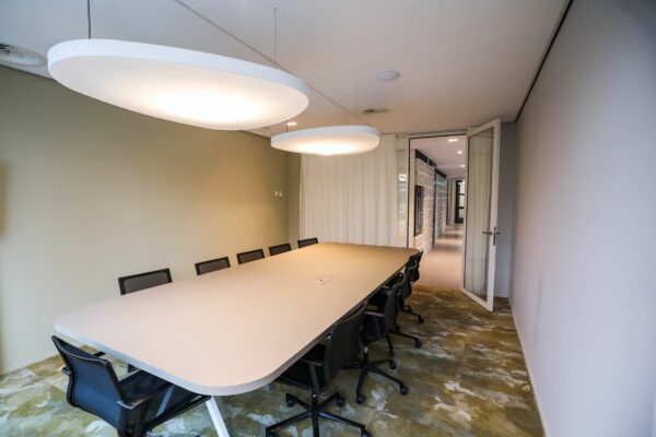 Vergaderruimte met lange tafel, tien stoelen, moderne verlichting en zicht op een gang door een open glazen deur.