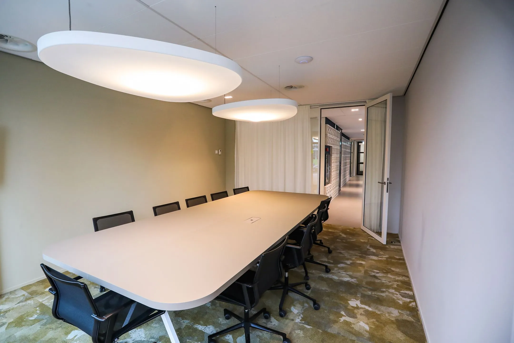 Vergaderruimte met lange tafel, tien stoelen, moderne verlichting en zicht op een gang door een open glazen deur.