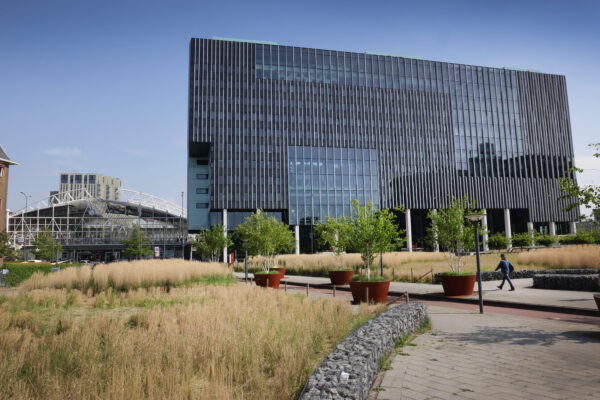 Een modern gebouw met glas en verticale lijnen, omgeven door groen en een wandelpad.