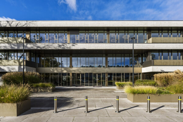 Modern kantoorgebouw met grote ramen en groene planten ervoor onder een blauwe hemel.