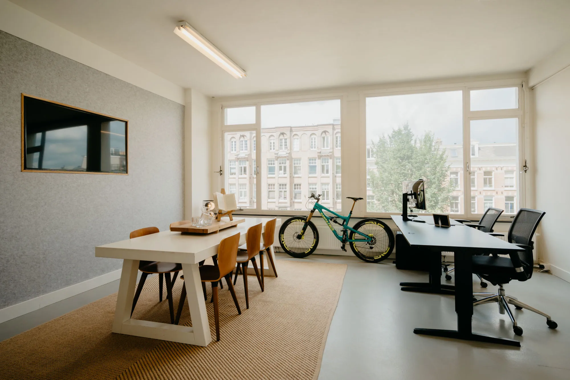 Een modern kantoor met een vergadertafel, fiets en bureau voor grote ramen met uitzicht op historische gebouwen.