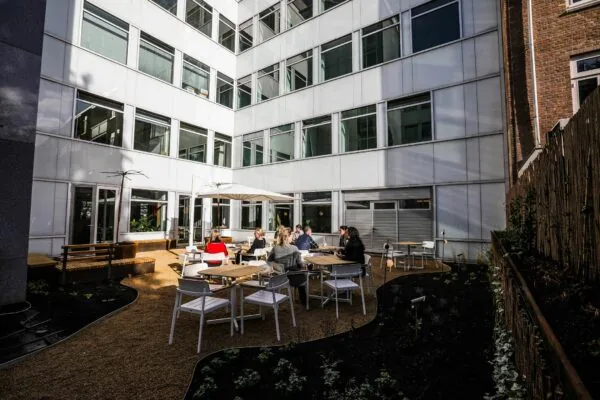 Mensen zitten en praten aan tafels in een zonnige binnentuin tussen kantoorgebouwen.