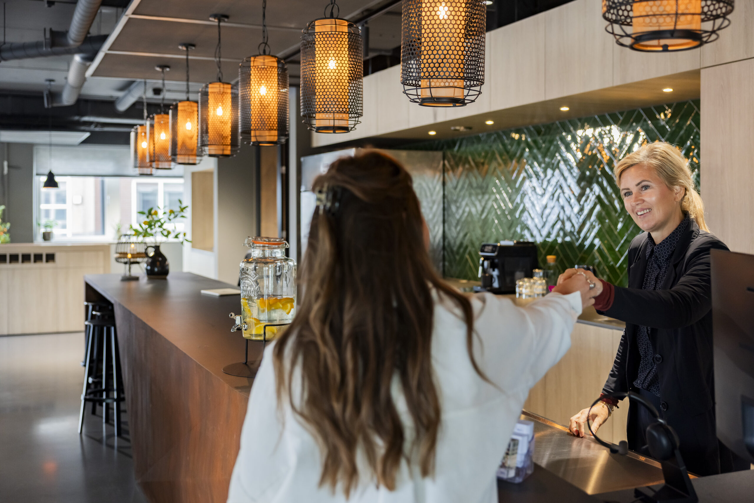 Een vrouw bij een elegante entreebalie begroet een bezoeker in een modern interieur met hanglampen en drankverfrissing.