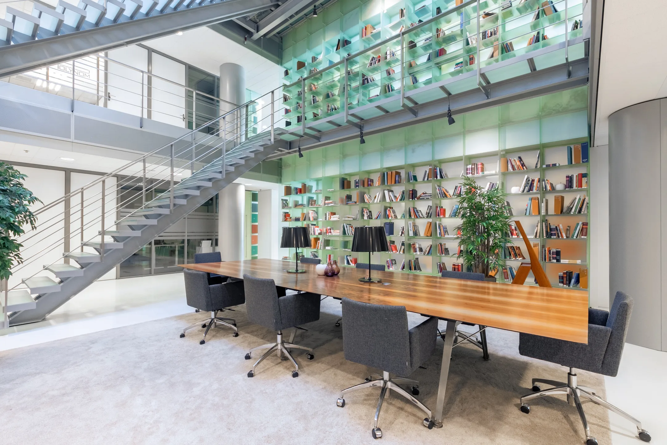 Een moderne bibliotheek met een grote houten tafel, grijze stoelen en een trap naast een muur met boekenplanken.