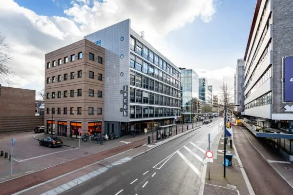 Straatbeeld met het kantoorgebouw 'Jacob' aan een brede weg met bushalte, winkels en moderne architectuur.