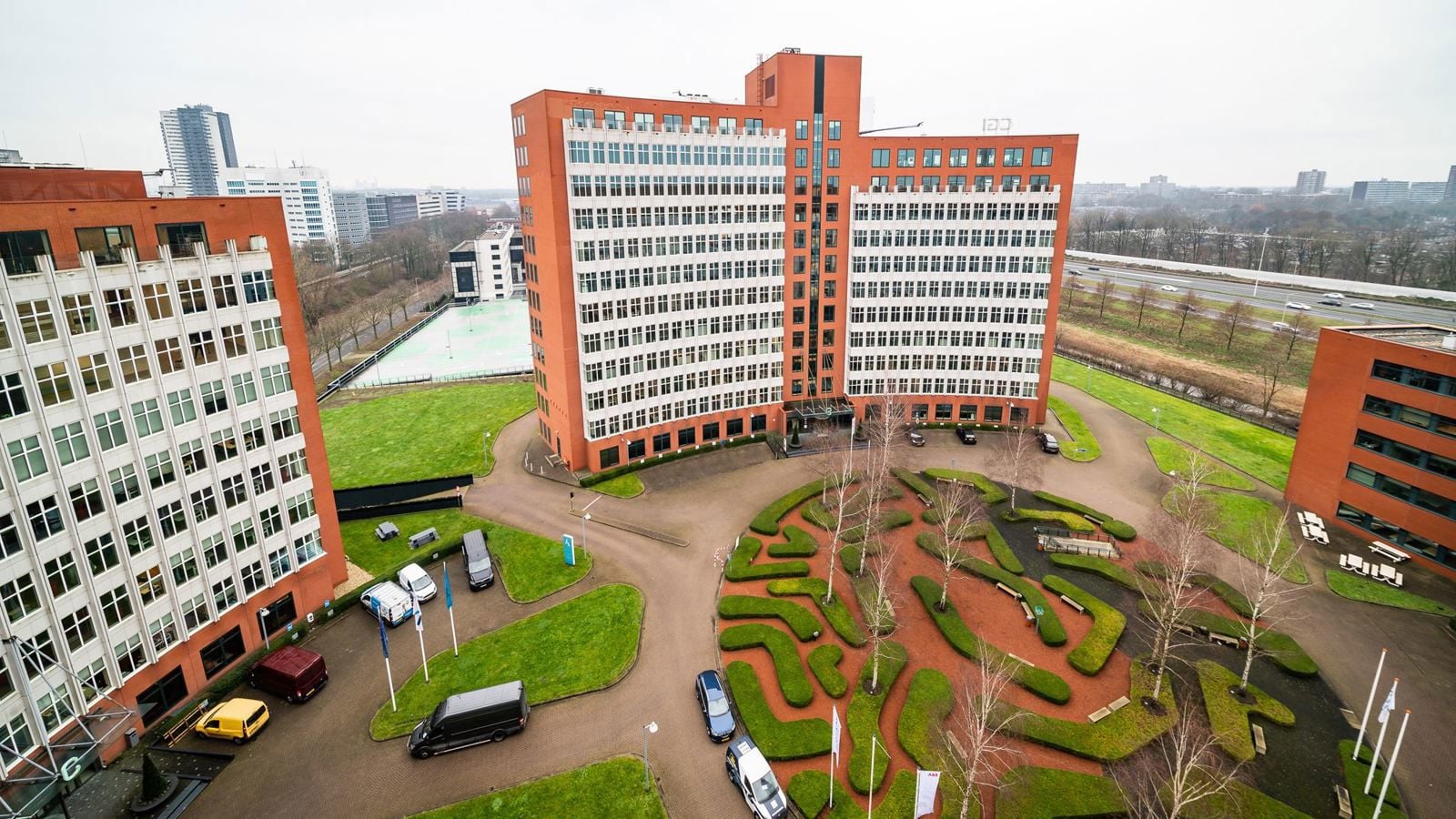 Luchtfoto van een groot kantoorgebouw met omliggende groenvoorzieningen en geparkeerde voertuigen.