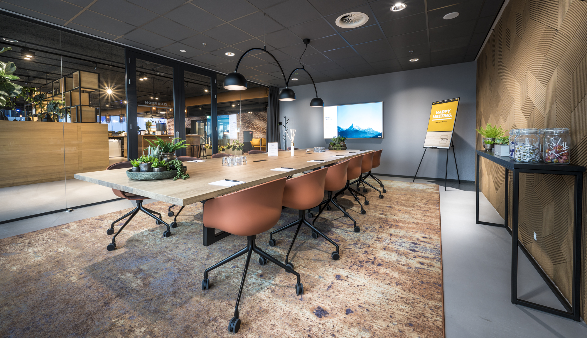 Een moderne vergaderruimte met een lange tafel, comfortabele stoelen, planten en een groot scherm aan de muur.
