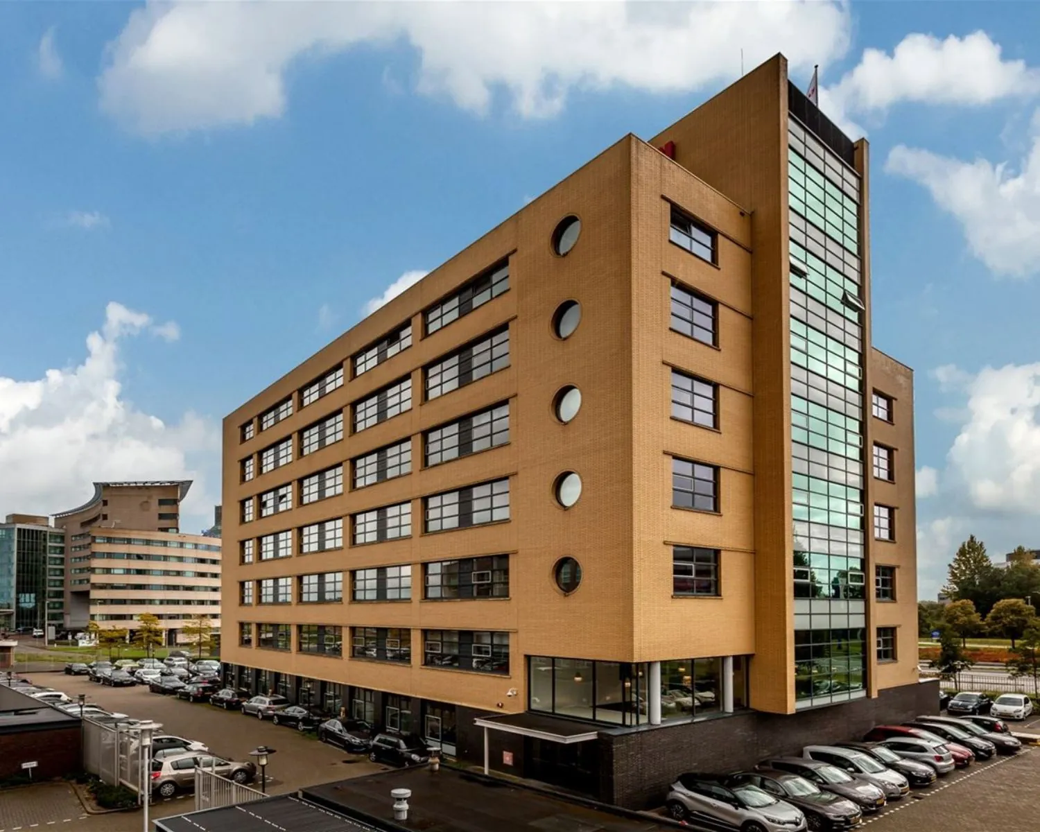 Modern kantoorgebouw met oranje bakstenen gevel en grote ramen, omringd door geparkeerde auto's.