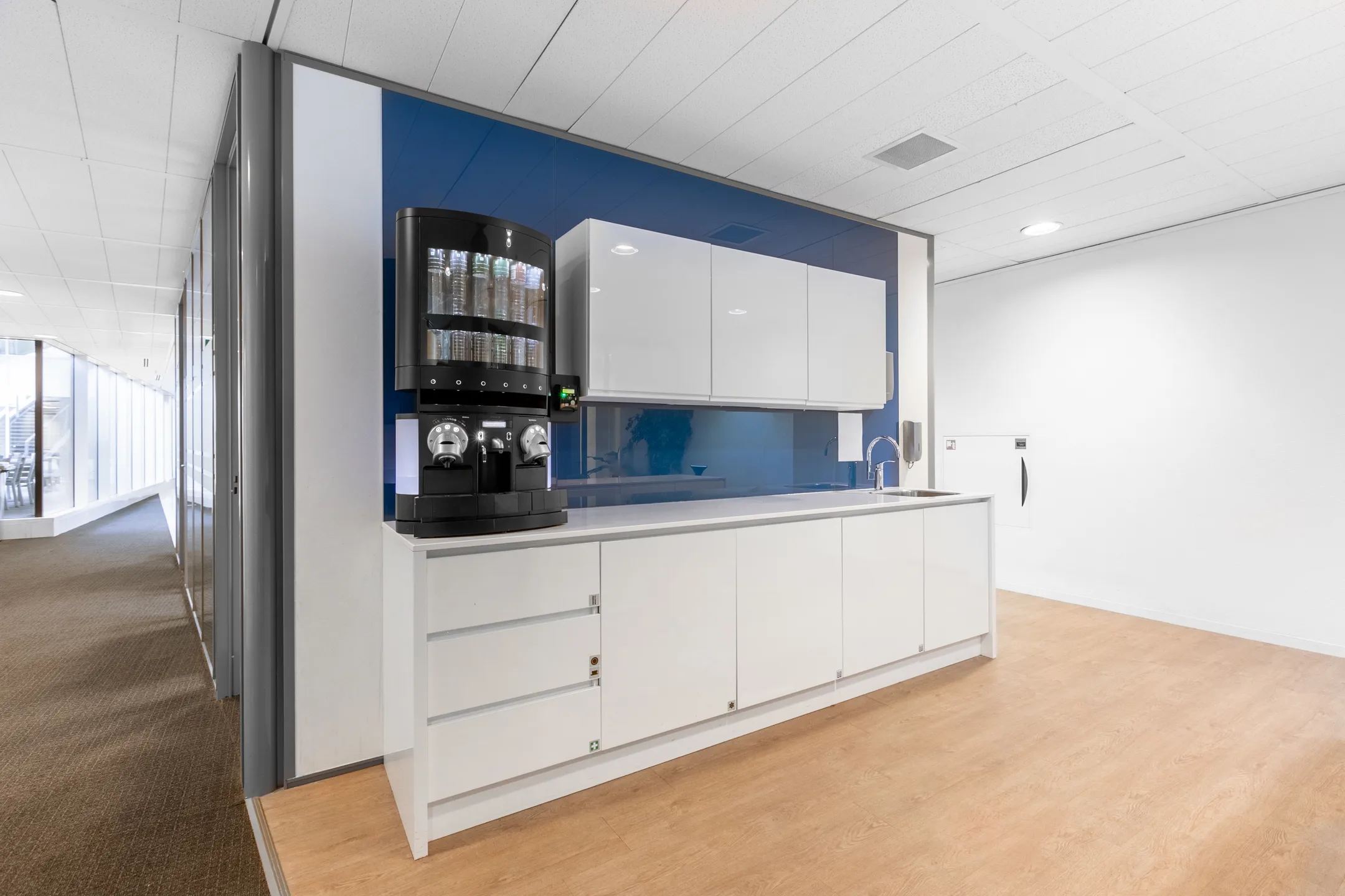 Een moderne kantoorruimte met een witte kitchenette en koffieapparaat tegen een blauwe wand.