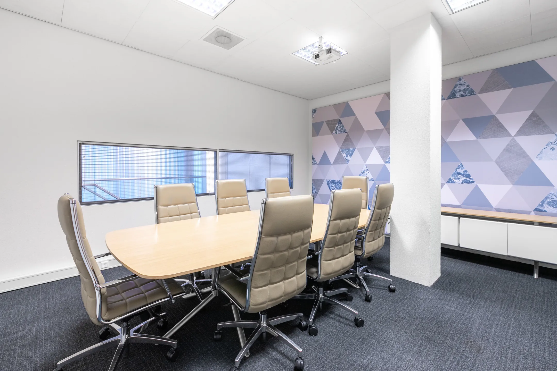 Vergaderruimte met beige stoelen rond een houten tafel en een wand met geometrisch patroon.