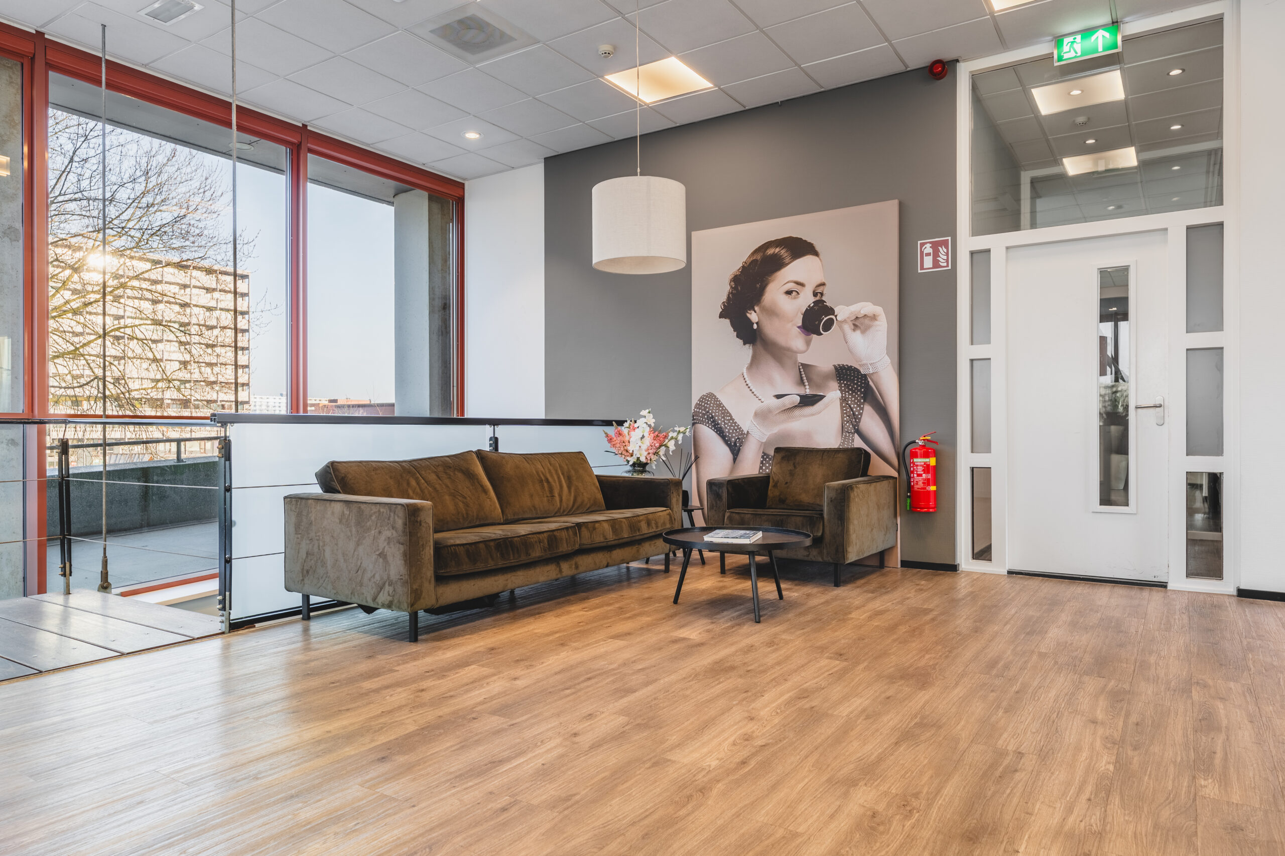 Een moderne kantoorruimte met een vintage poster van een vrouw die thee drinkt, omgeven door een bruine bank en fauteuil.