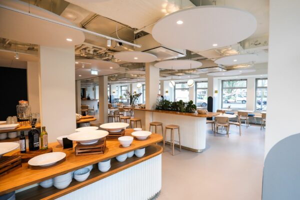 Moderne, open bedrijfskantine met houten meubels, veel lichtinval en een buffetopstelling met witte schalen.