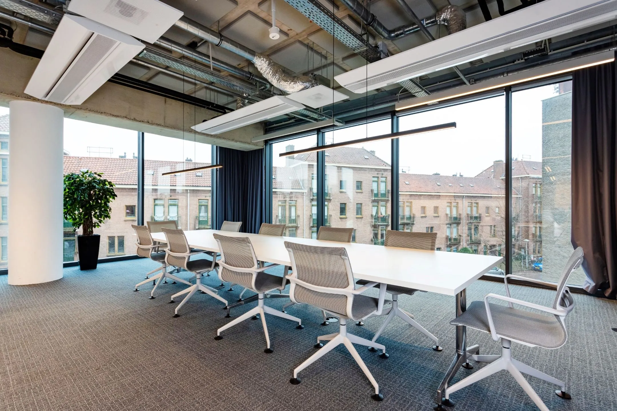 Moderne vergaderruimte met een lange witte tafel, grijze bureaustoelen en grote ramen met uitzicht op een bakstenen woongebouw.
