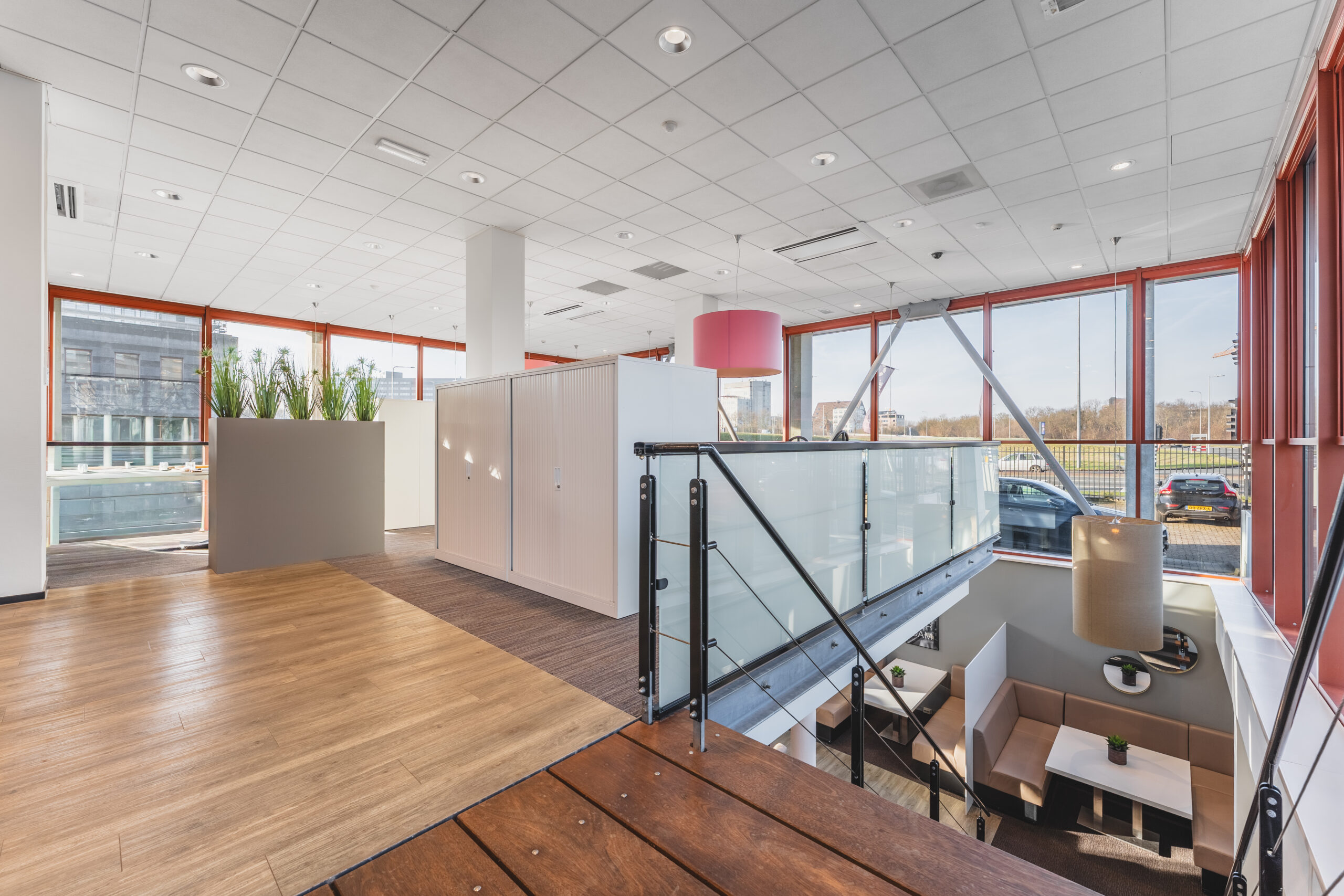 Een moderne kantoorruimte met glazen balustrade, houten vloer en grote ramen die veel natuurlijk licht binnenlaten.