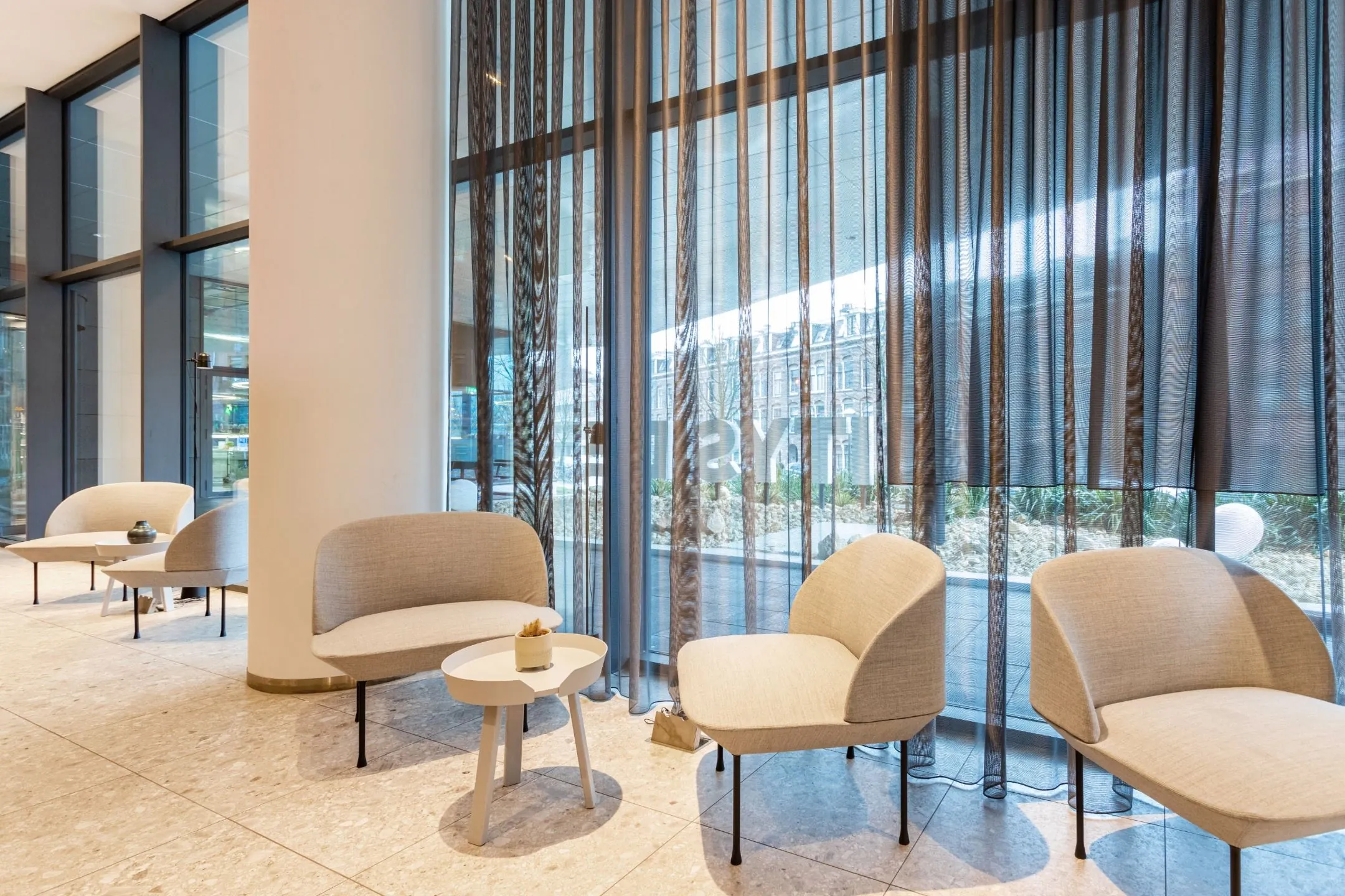 Moderne wachtruimte met beige stoelen, kleine ronde tafels en hoge ramen met transparante gordijnen.