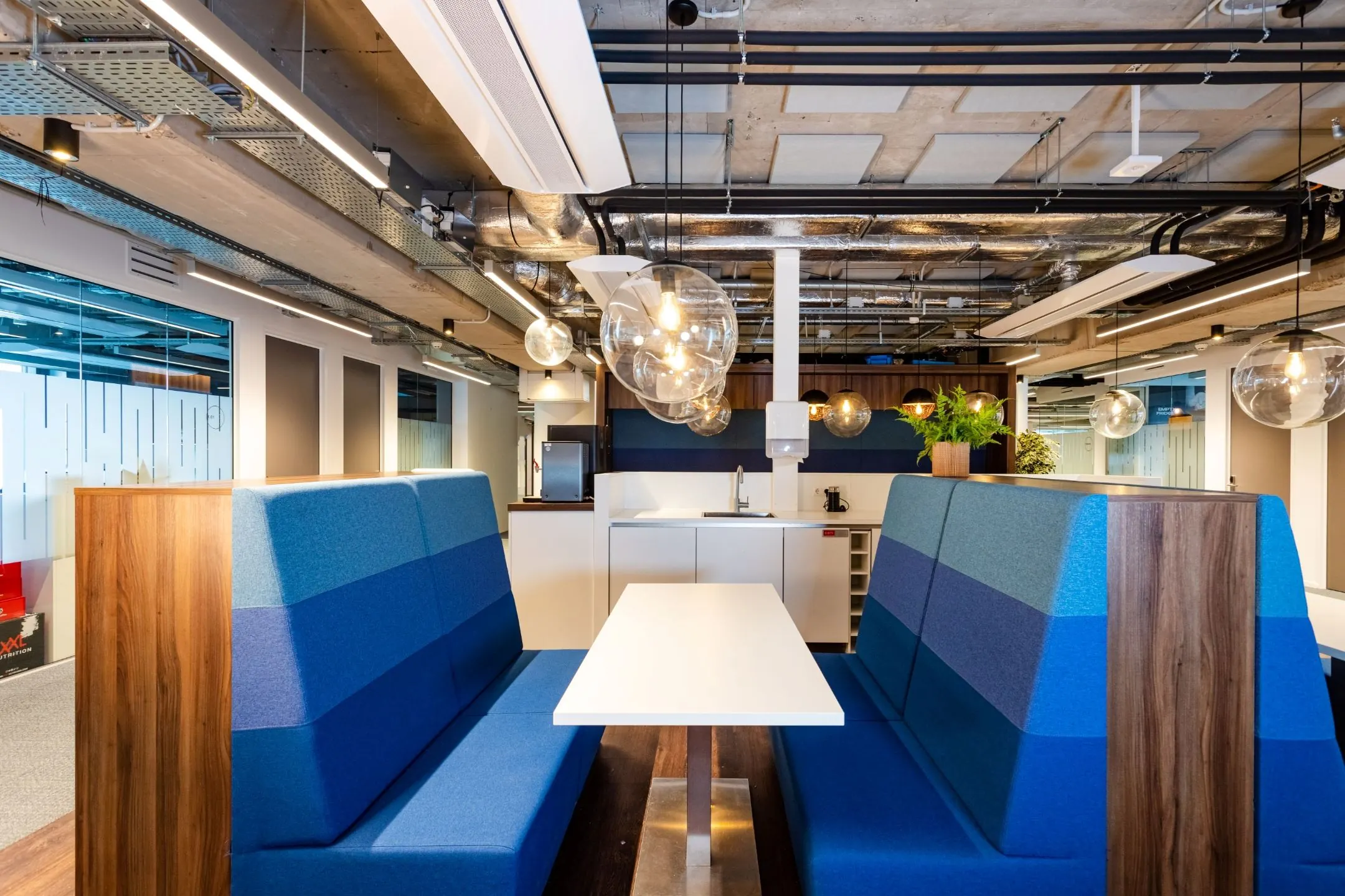 Moderne kantoorruimte met blauwe zitbanken, een witte tafel en een open kitchenette met industriële plafondafwerking.
