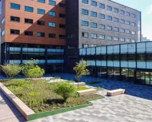 Binnenplaats met moderne bestrating, plantenbakken, zitbanken en omringende bakstenen en glazen gebouwen.