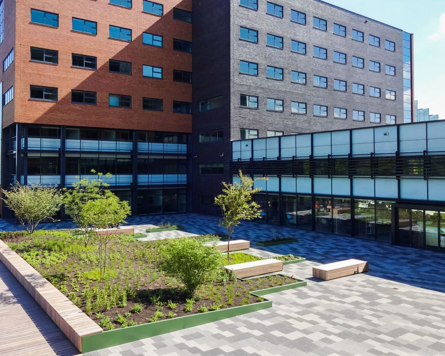 Binnenplaats met moderne bestrating, plantenbakken, zitbanken en omringende bakstenen en glazen gebouwen.