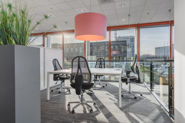 Een moderne kantoorruimte met bureaus, zwart gestoffeerde stoelen, een grote roze hanglamp en veel daglicht.