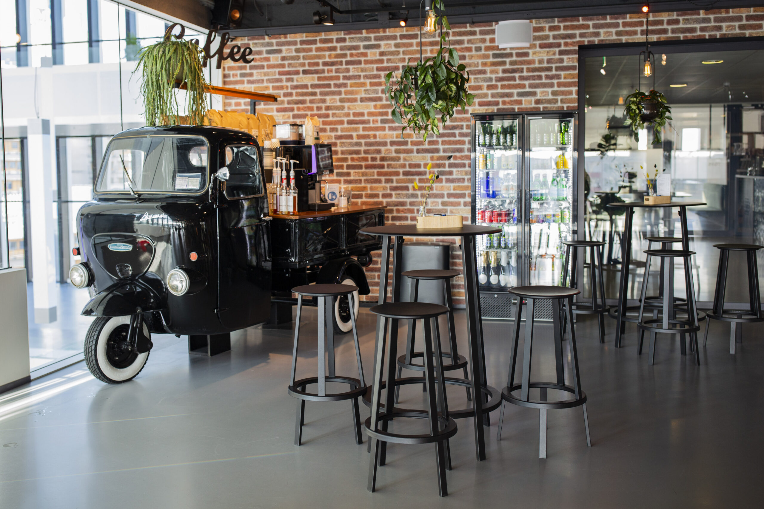 Koffiehoek met een zwarte driewieler als toonbank, hoge tafels en stoelen, en een muur van bakstenen.