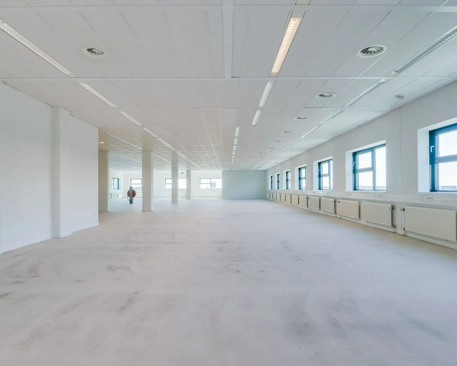 Ruime, lichte kantoorruimte met witte muren, een systeemplafond en grote ramen aan één zijde.