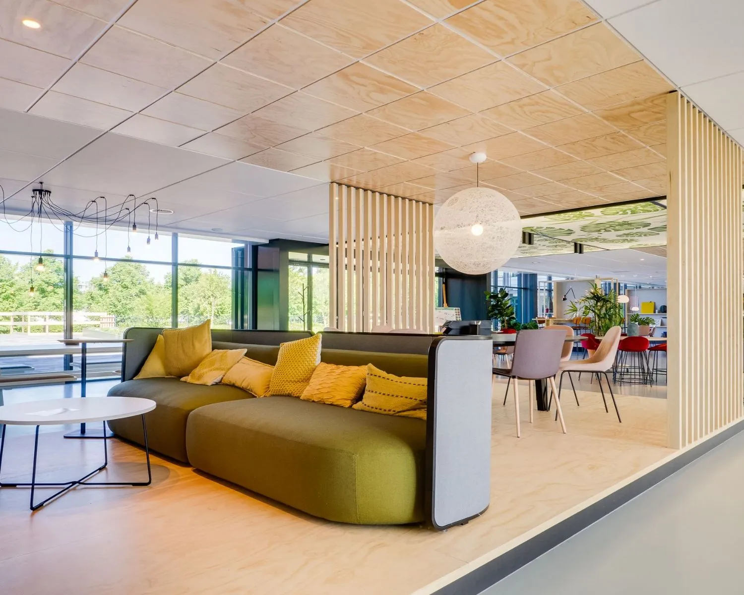 Moderne en kleurrijke kantoorruimte met een groene bank, houten details en grote ramen die uitzicht bieden op een groene omgeving.