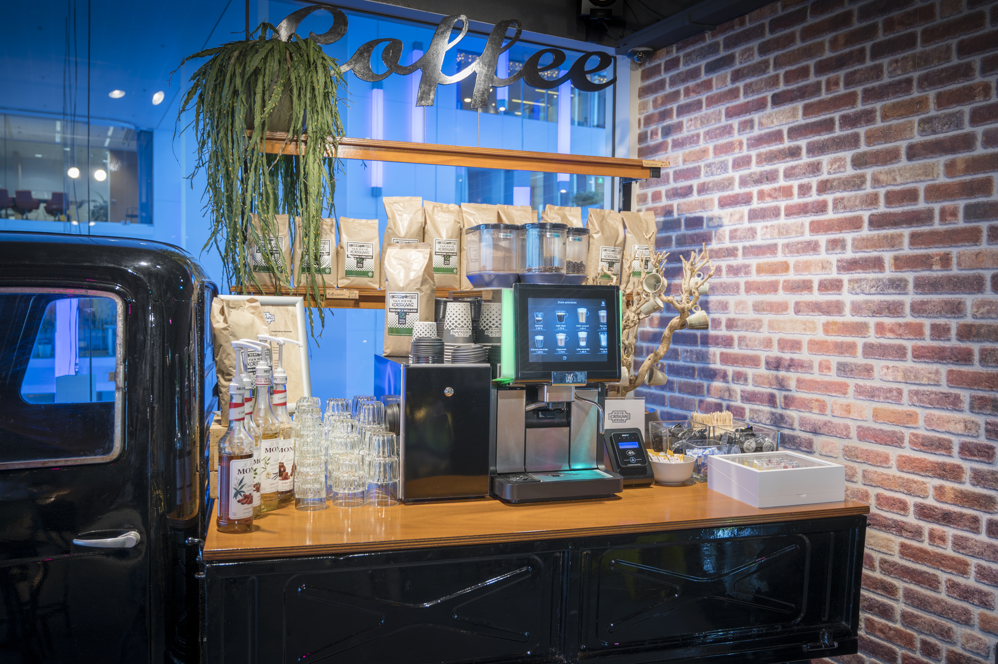 Een sfeervolle koffiestand met een espressomachine, siropen en koffiezakken, naast een muur met baksteenmotief.