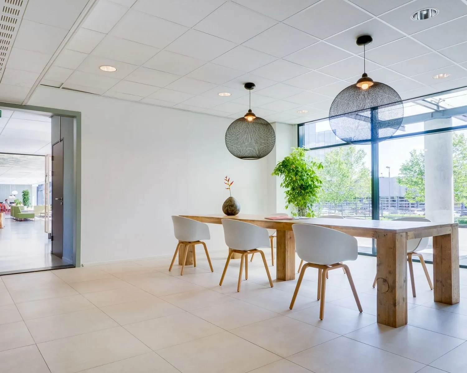 Moderne en lichte kantoorlounge met houten tafel, witte stoelen en grote ramen met uitzicht op groen.