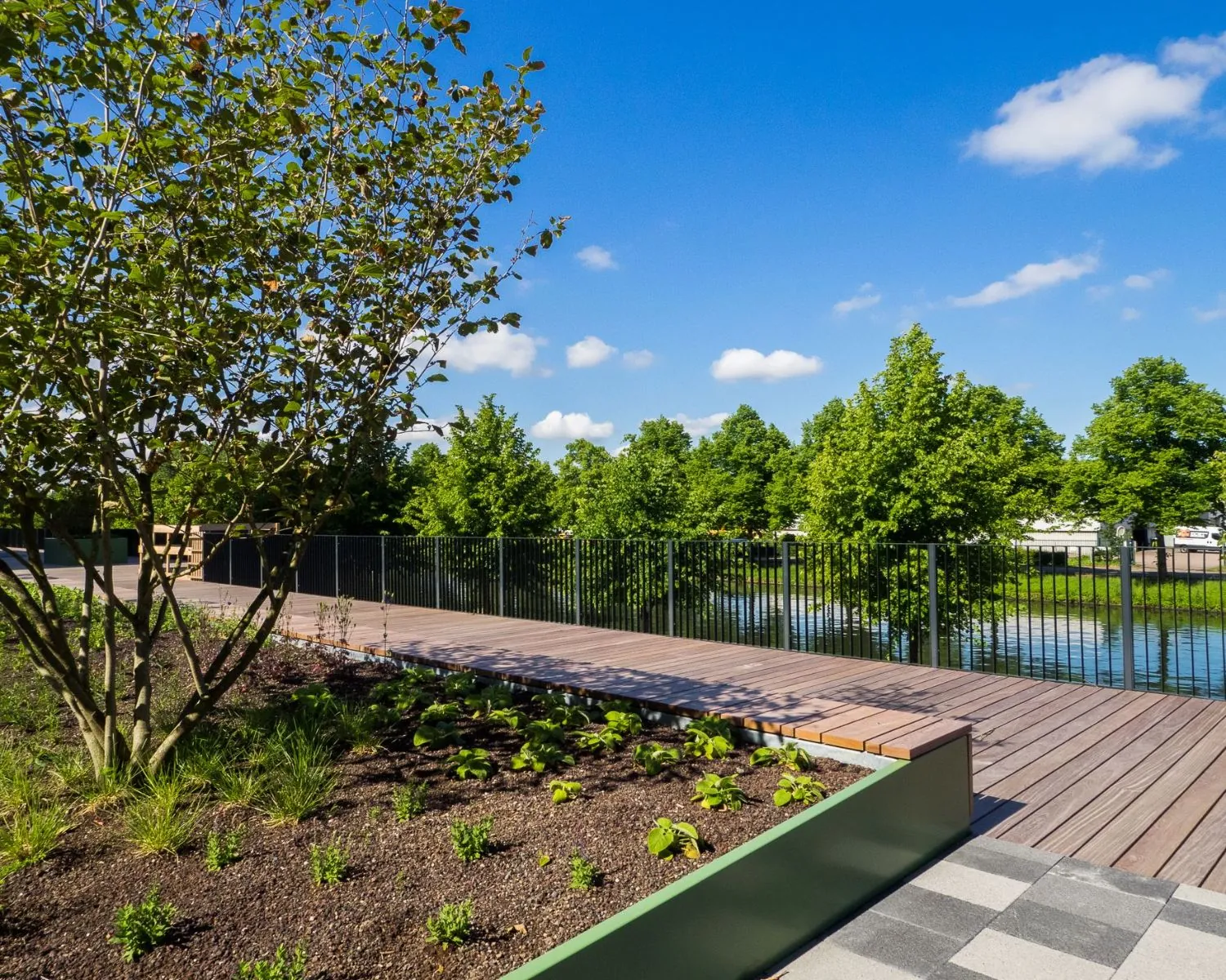 Houten wandelpad met zwart hek langs een waterpartij, omgeven door groene bomen en een aangelegde tuin.