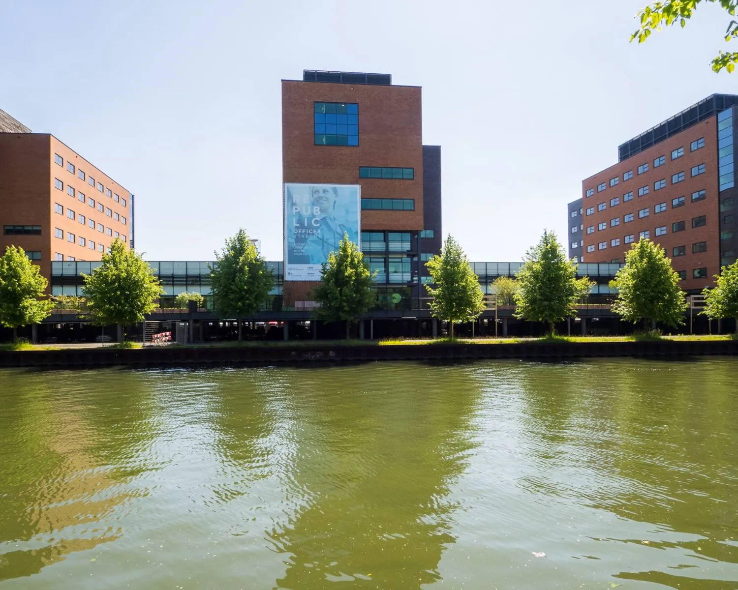 Kantoorgebouw 'The Re-.Public Offices' met bakstenen gevels aan het water, omgeven door bomen.