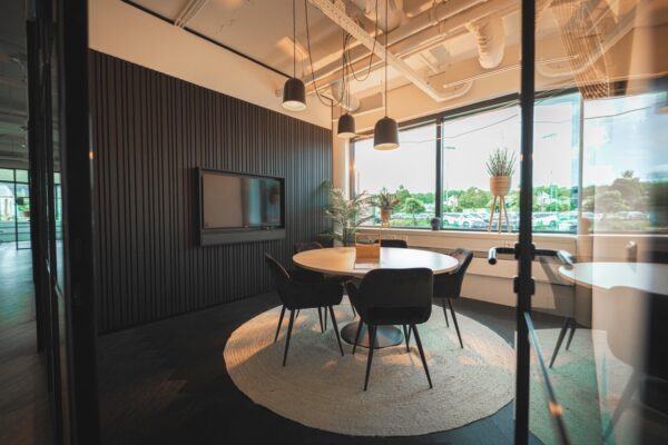 Moderne vergaderruimte met ronde tafel, vijf stoelen, een tv-scherm en grote ramen met uitzicht op een parkeerterrein.