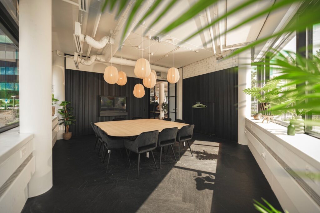 Moderne vergaderruimte met houten tafel, zwarte stoelen, hanglampen en veel natuurlijk licht.
