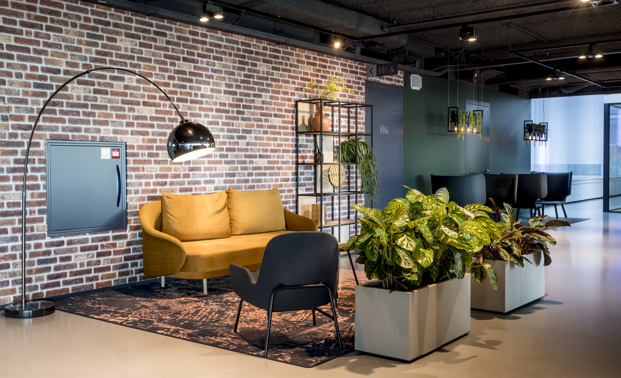 Een sfeervolle zithoek met een gele bank, een zwarte stoel, en planten tegen een muur van baksteen.