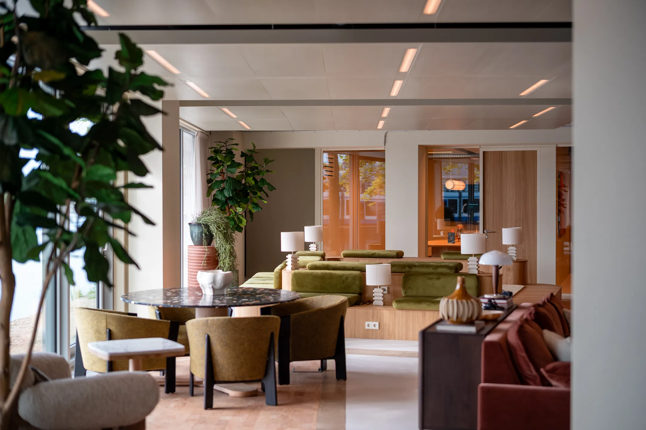 Een moderne woonkamer met groene zitbanken, ronde tafel, stoelen en diverse kamerplanten.