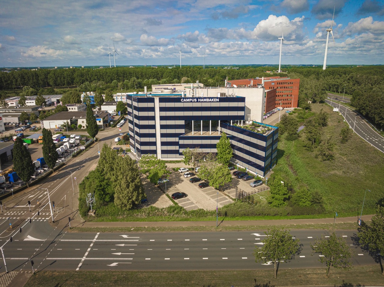 Luchtfoto van het moderne kantoorgebouw Campus Hambaken, omgeven door groen, wegen en windmolens in de achtergrond.
