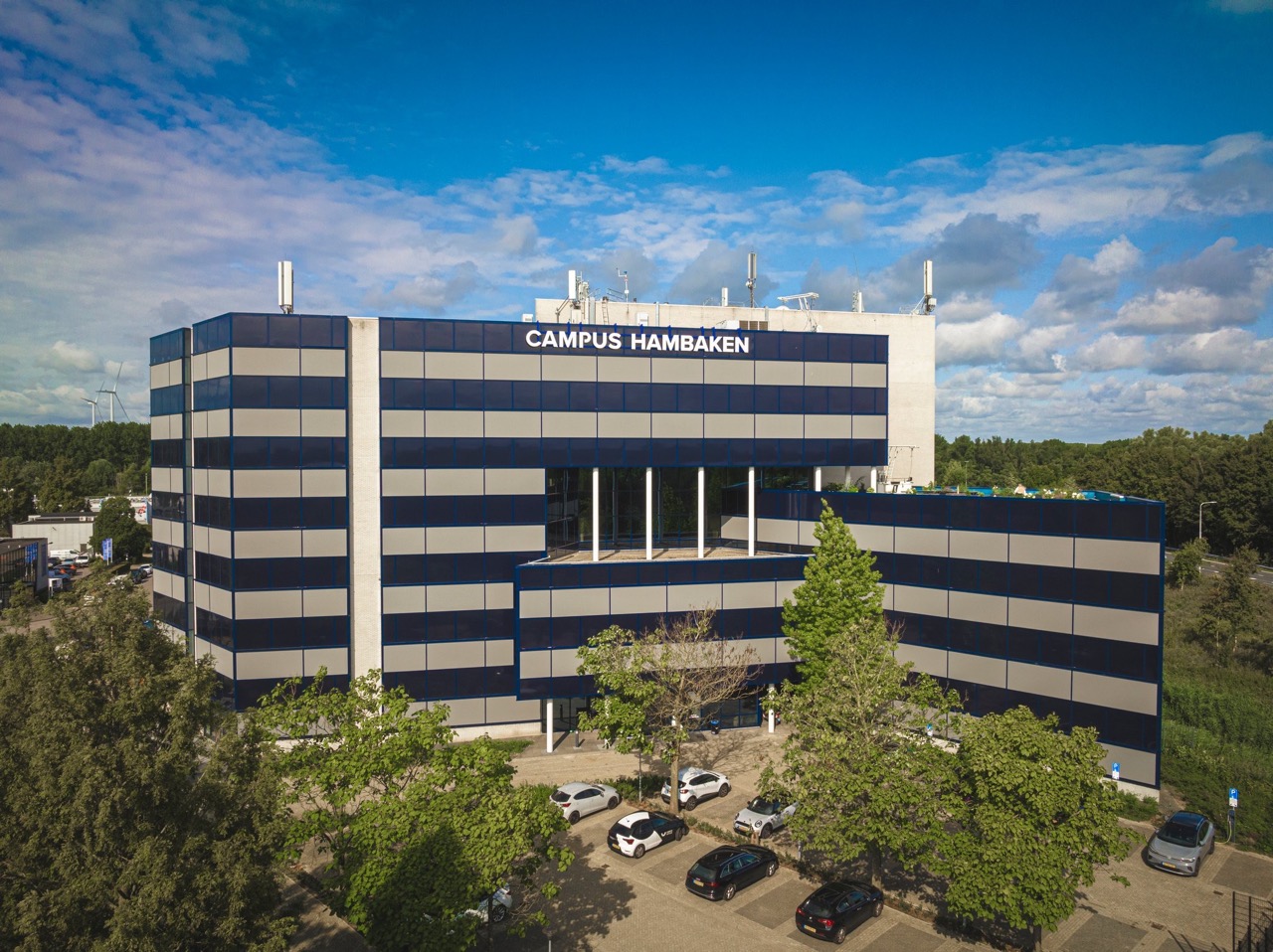 Modern kantoorgebouw Campus Hambaken met blauwe en grijze gevelpanelen, omringd door bomen en geparkeerde auto's.
