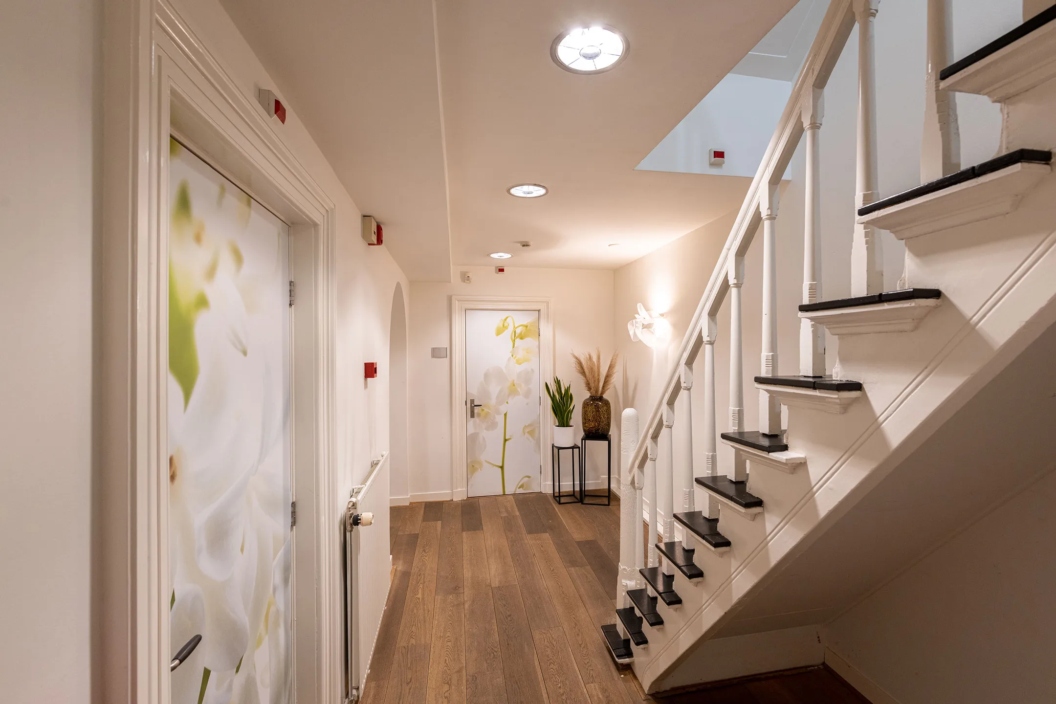 Een lichte hal met een houten vloer, een witte trap, en deuren met bloemenafbeeldingen.