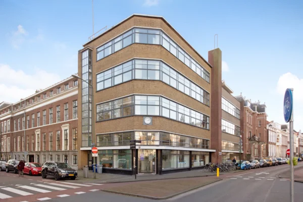 Een hoekpand met een modernistische uitstraling en grote ramen aan een straat met geparkeerde auto's en enkele fietsers.