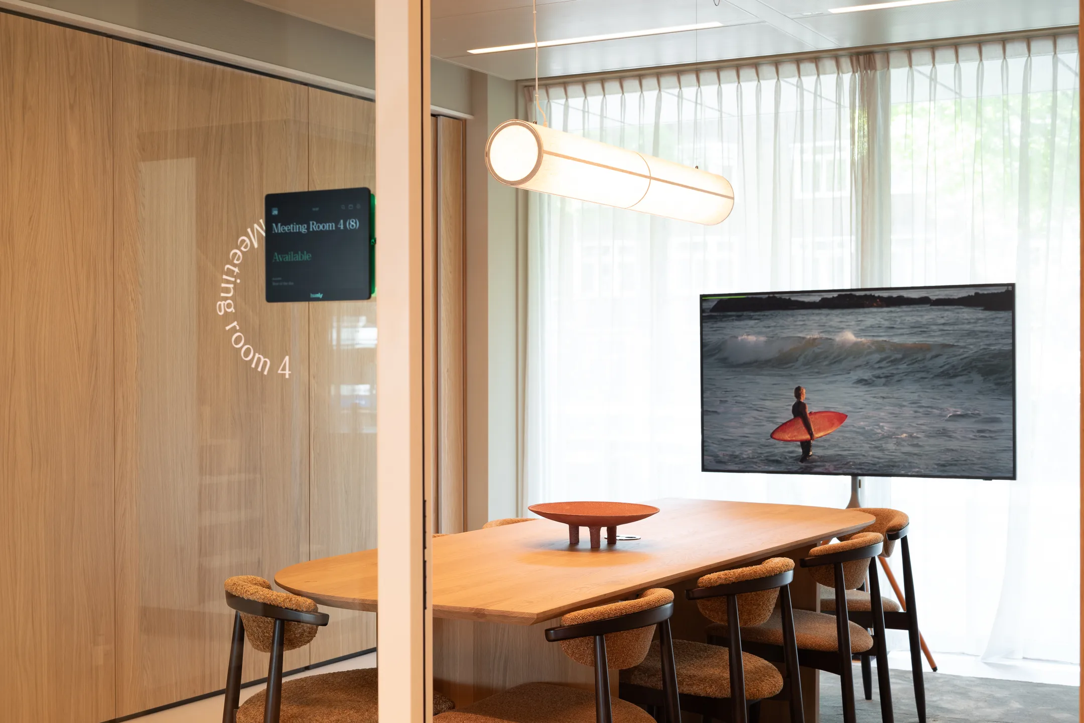 Een moderne vergaderruimte met een tafel, stoelen, scherm met een surfer en een bord op de deur met "Meeting Room 4".