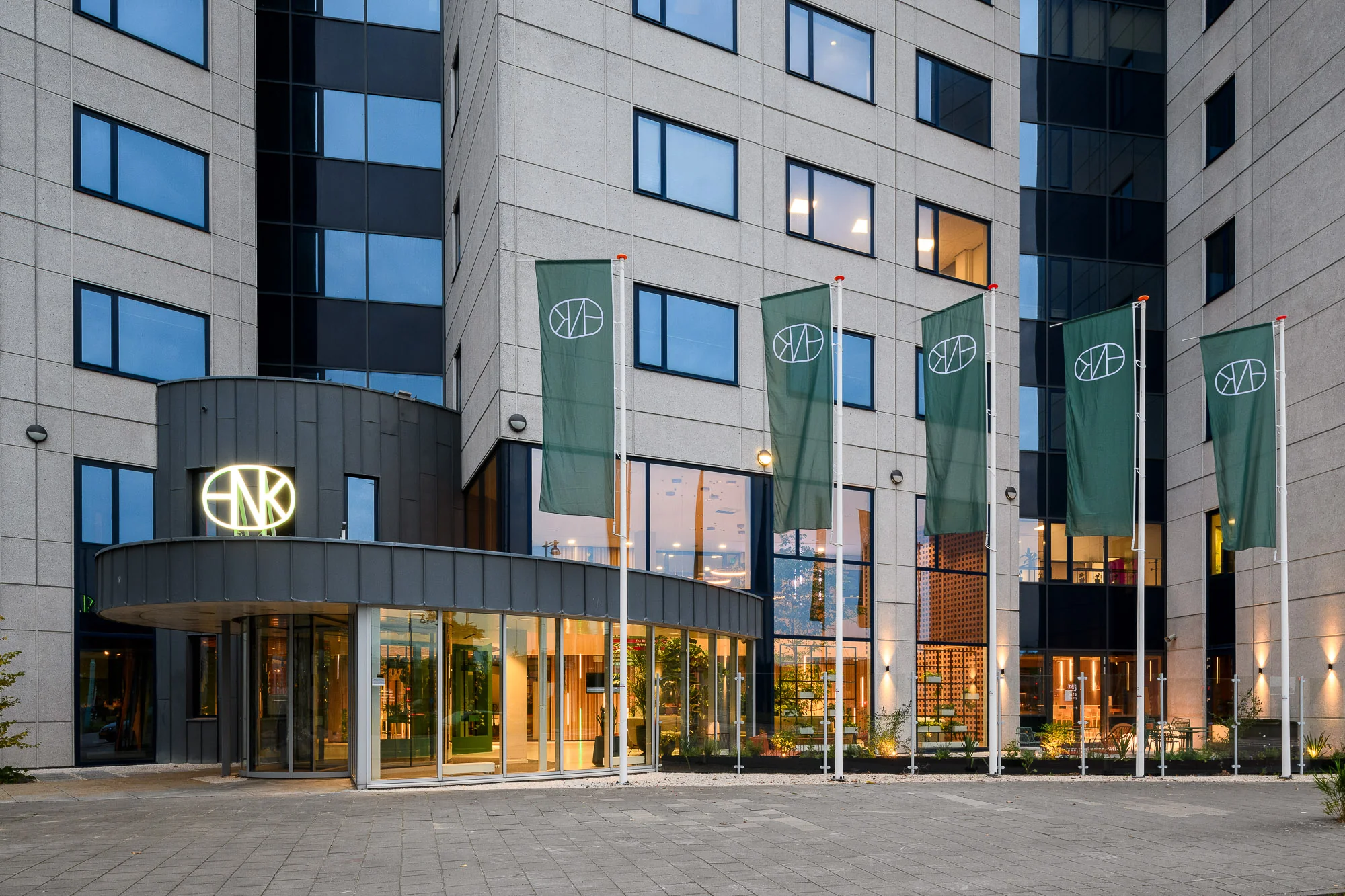 Voorgevel van HNK Amsterdam Sloterdijk met groene vlaggen en verlichte entree.
