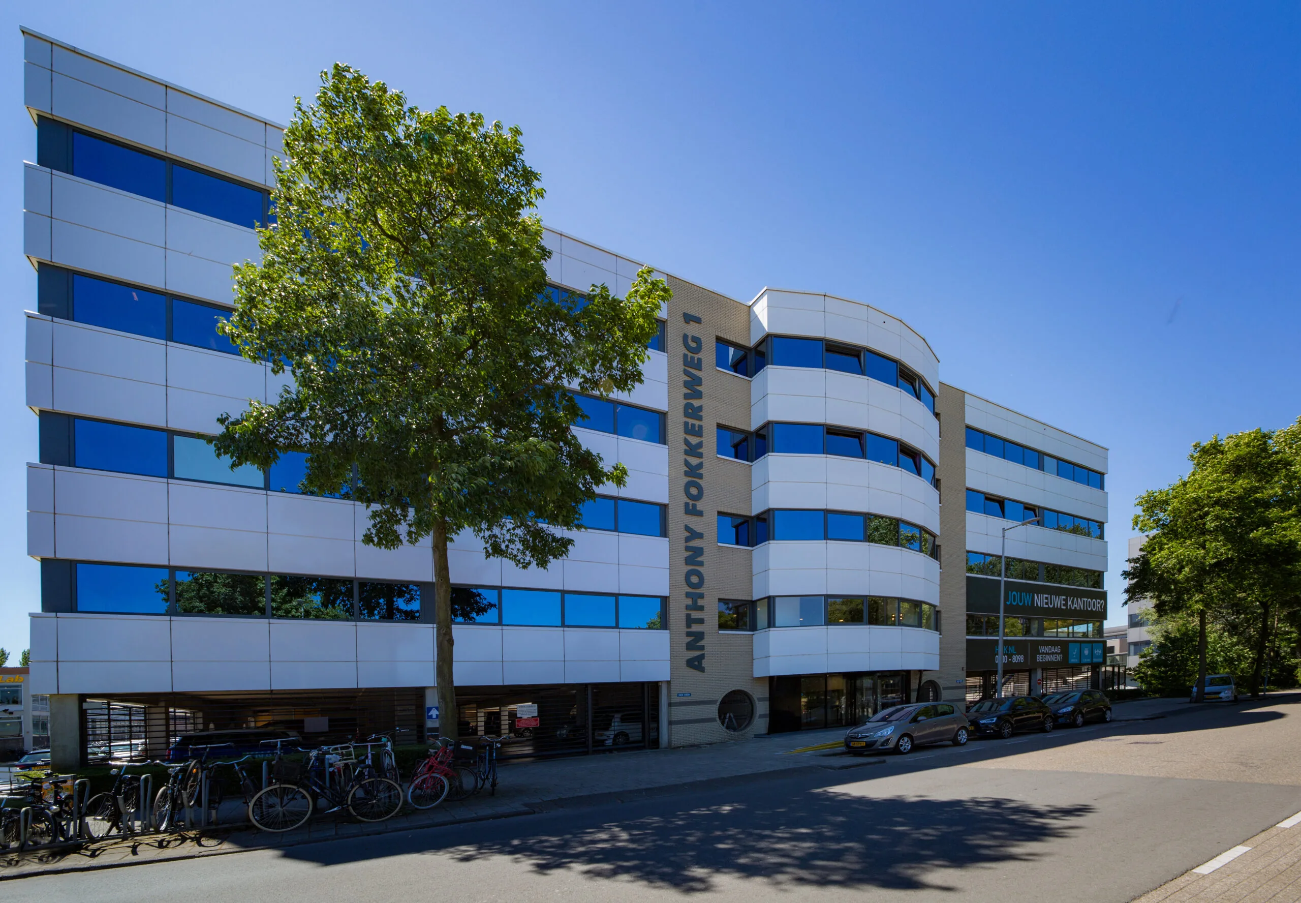 Modern kantoorgebouw HNK Amsterdam Schinkel aan de Anthony Fokkerweg 1 met fietsenstalling en geparkeerde auto’s voor de ingang.