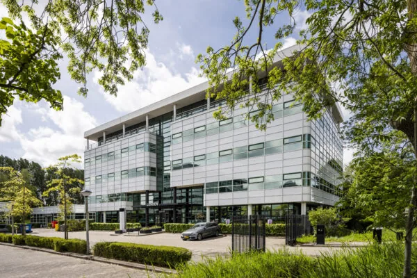 Modern kantoorgebouw HNK Amsterdam Zuidoost, omgeven door groen en met parkeergelegenheid voor de ingang.