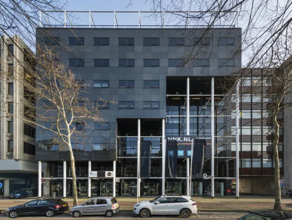 Voorgevel van het HNK-gebouw in Rotterdam Centrum met glazen entree en geparkeerde auto's op de voorgrond.
