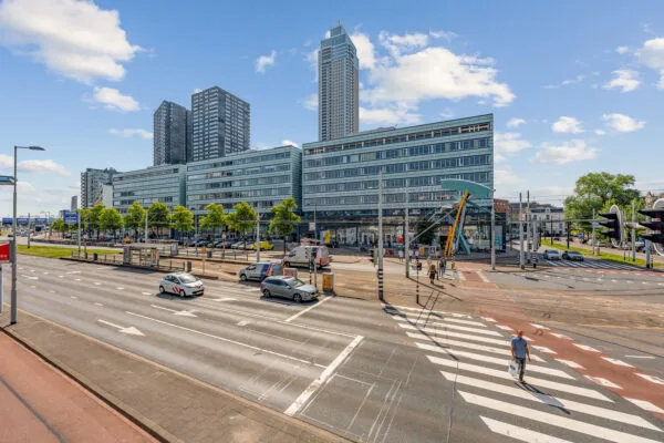 Drukke stadsweg met zebrapad, moderne hoogbouw en een glazen kantoorcomplex op een zonnige dag.