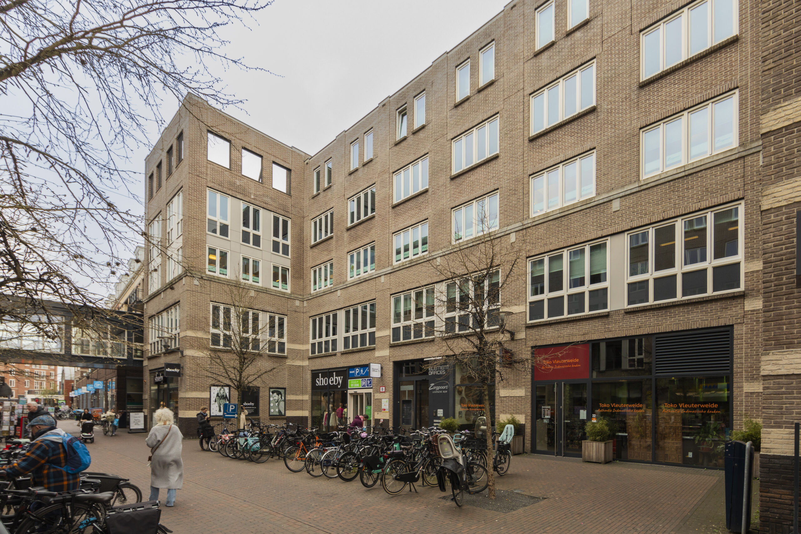 Een modern bakstenen gebouw met winkels op de begane grond in Vleuterweide, omringd door geparkeerde fietsen en voorbijgangers.