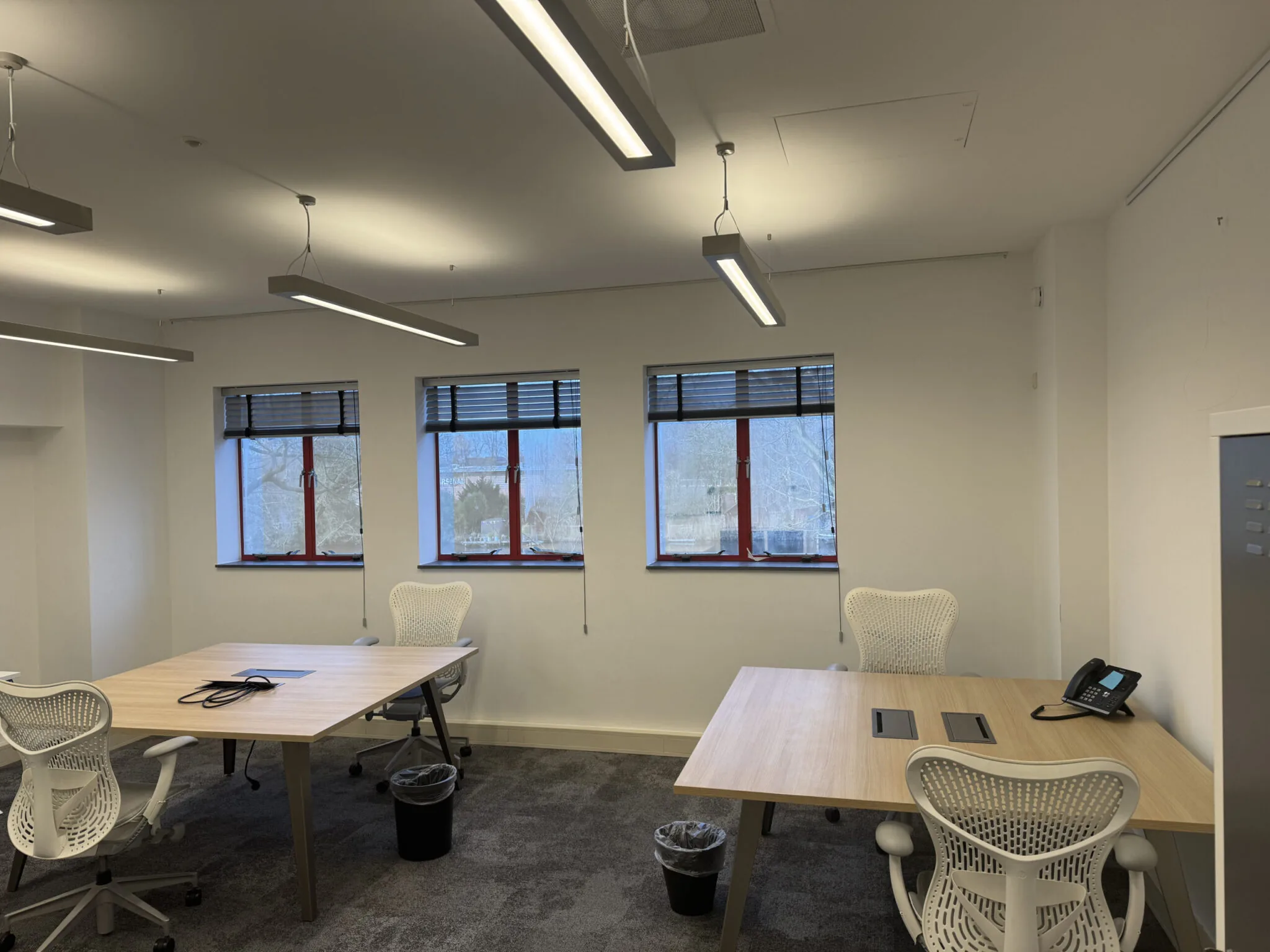 Modern kantoorruimte 1.01D met bureaus, ergonomische stoelen, telefoontoestel en drie ramen met jaloezieën.