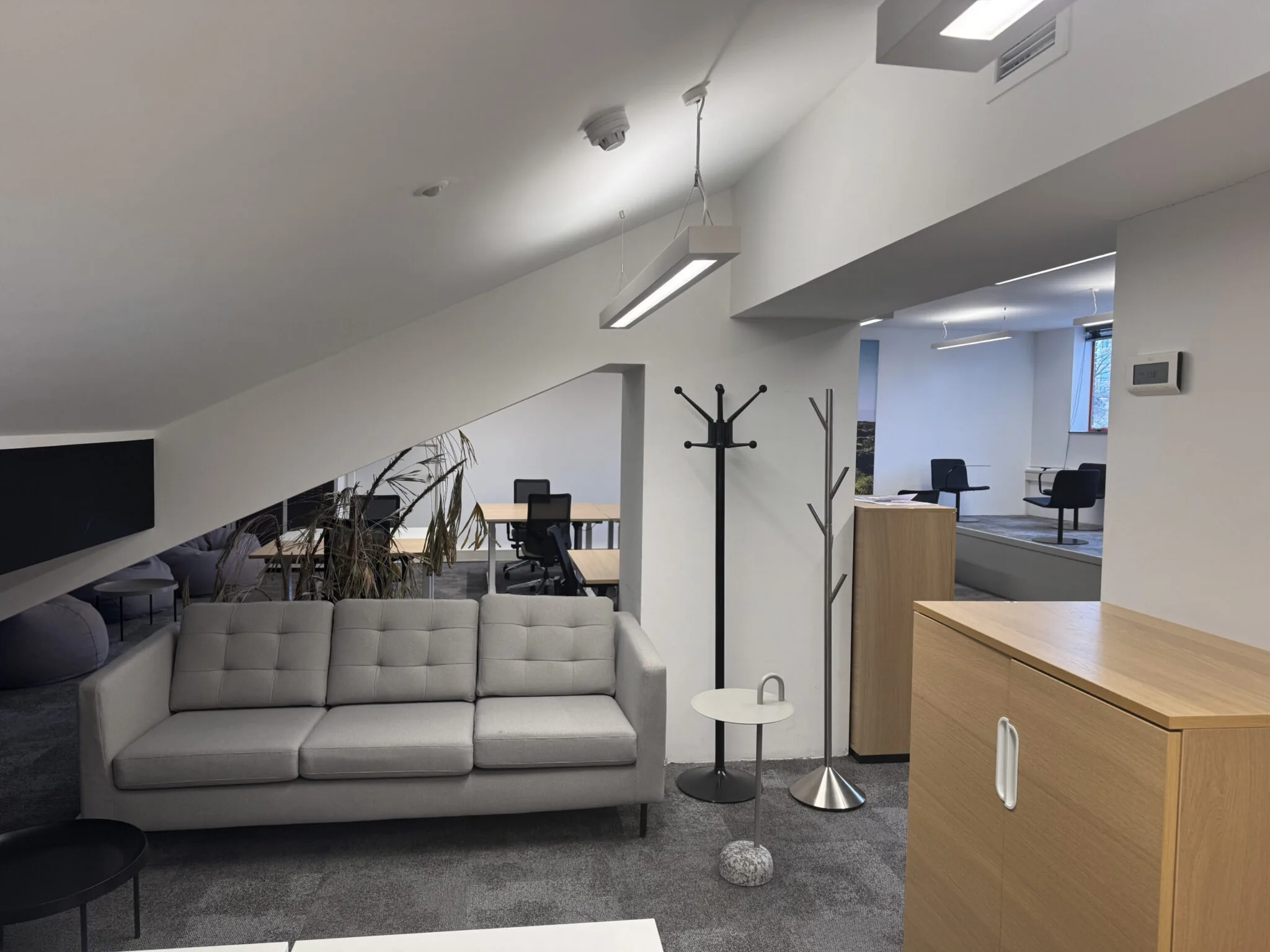 Moderne zitruimte in een kantoor met een grijze bank, kapstokken, tafeltjes en werkplekken op de achtergrond.